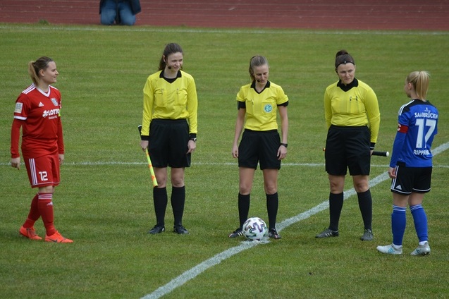 Zwei Fu&szlig;ballspielerinnen und vier Schiedsrichterinnen stehen auf dem Spielfeld zur Spielanpfiff bereit.