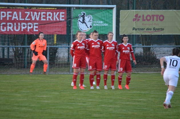 F&uuml;nf Fu&szlig;ballspielerinnen in roten Trikots bereiten sich auf einen Freisto&szlig; vor, w&auml;hrend eine Torh&uuml;terin im Hintergrund steht.