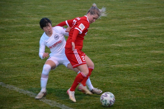 Zweikampf zwischen zwei Fu&szlig;ballspielerinnen auf dem Rasen, eine in roter und eine in wei&szlig;er Uniform.