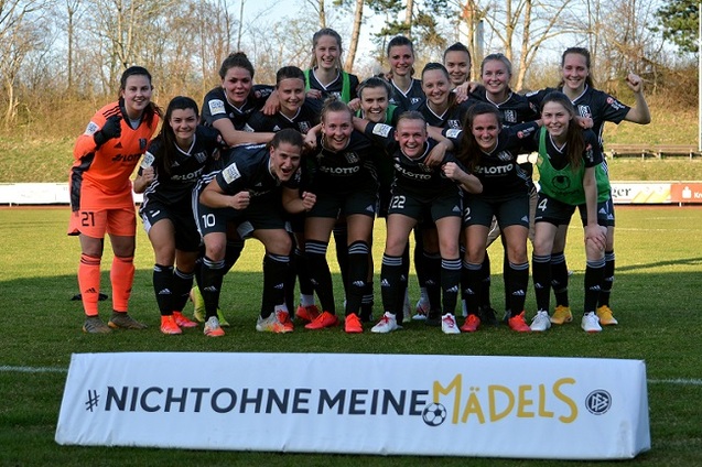 Gruppenfoto einer Frauenfu&szlig;ballmannschaft in schwarzen Trikots, posierend vor einem Banner mit dem Text &bdquo;NICHT OHNE MEINE M&Auml;DELS&ldquo;.