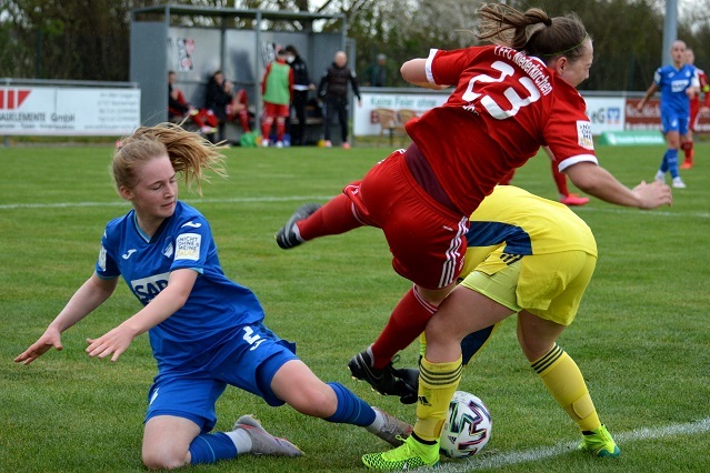 Zwei Fu&szlig;ballspielerinnen k&auml;mpfen um den Ball, w&auml;hrend eine dritte Torh&uuml;terin im Hintergrund positioniert ist.