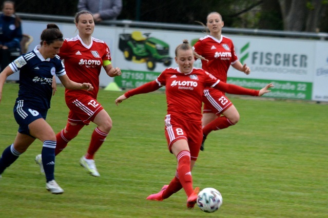 Vier Frauen im Fu&szlig;balltrikot stehen auf einem Rasenfeld; eine Spielerin schie&szlig;t einen Ball in Richtung Tor.