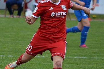 Spielerin in rotem Trikot tritt den Fu&szlig;ball auf einem Rasenfeld, w&auml;hrend eine Mitspielerin im blauen Trikot im Hintergrund steht.