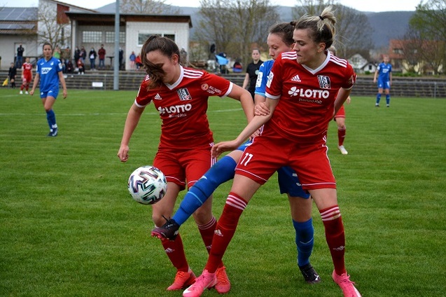 Zwei Fu&szlig;ballspielerinnen in roten Trikots k&auml;mpfen um den Ball auf einem gr&uuml;nen Spielfeld. Zuschauer im Hintergrund.