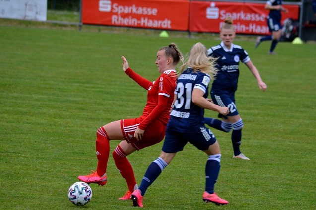 Zwei Fu&szlig;ballspielerinnen im Wettkampf um den Ball auf einem Rasenfeld, eine in rotem und eine in dunklem Trikot.