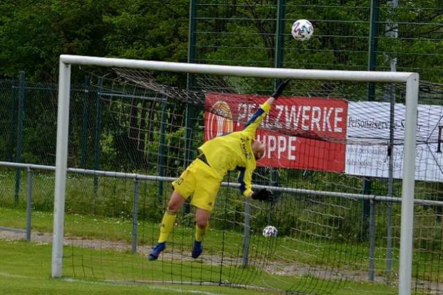 Torwart in gelber Uniform springt hoch, um einen Fu&szlig;ball in einem Tor zu fangen, Hintergrund mit Zaun und Werbebannern.