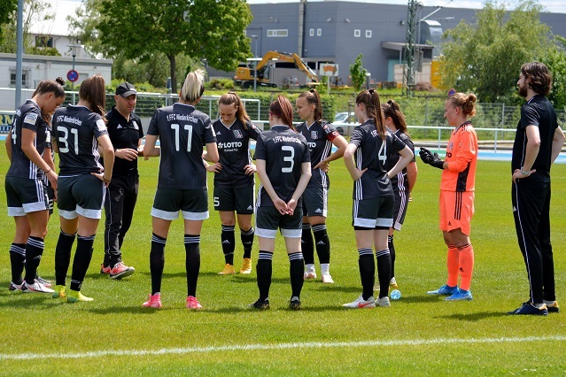 Trainer bespricht Taktik mit einer Frauenfu&szlig;ballmannschaft auf dem Platz, Spielerinnen versammelt in schwarzer Sportkleidung.