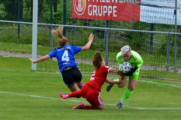 Torwartin in gr&uuml;n f&auml;ngt den Ball, w&auml;hrend eine Spielerin in rot und eine in blau am Boden liegen.
