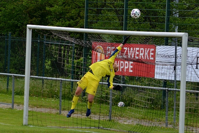 Torwart in gelber Uniform springt hoch, um einen Fu&szlig;ball in einem Tor zu fangen, Hintergrund mit Zaun und Werbebannern.