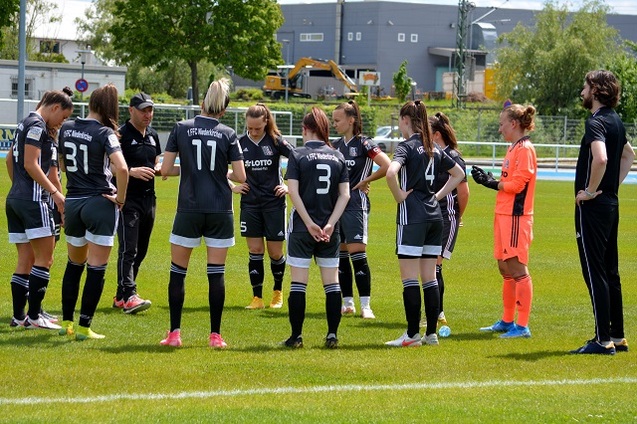 Trainer bespricht Taktik mit einer Frauenfu&szlig;ballmannschaft auf dem Platz, Spielerinnen versammelt in schwarzer Sportkleidung.