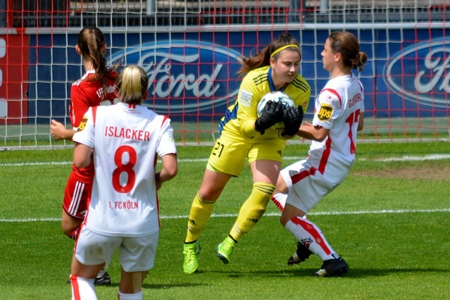 Torh&uuml;terin in gelbem Trikot f&auml;ngt den Ball vor zwei gegnerischen Spielerinnen auf dem Fu&szlig;ballfeld.
