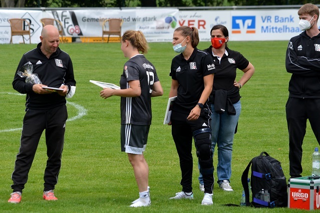 Zwei Trainer und drei Spielerinnen stehen auf einem Fu&szlig;ballfeld und tauschen Zertifikate bei einer &Uuml;bergabe.