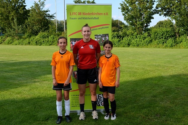 Drei M&auml;dchen in Fu&szlig;balltrikots stehen vor einem Vereinsbanner auf einem Sportplatz.