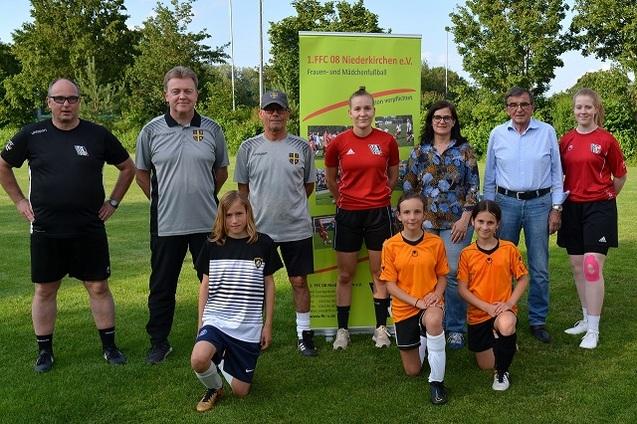 Gruppenbild mit Trainern und Spielerinnen des 1. FFC 08 Niederkirchen auf einem Fu&szlig;ballfeld vor einem Vereinsschild.