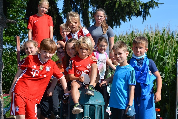 Eine Gruppe von Kindern posiert fr&ouml;hlich auf einem alten Traktor vor einem Maisfeld bei sonnigem Wetter.