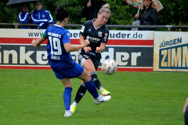 Zwei Fu&szlig;ballspielerinnen im Wettkampf um den Ball auf einem Rasenplatz, Zuschauer im Hintergrund mit Regenschirmen.