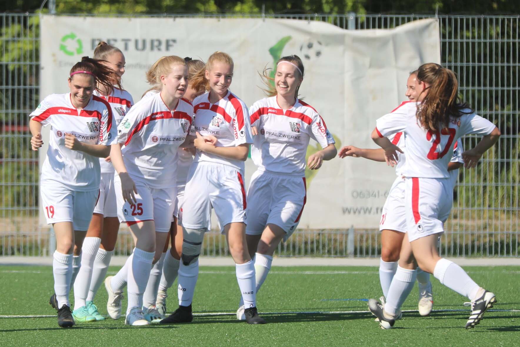 Gruppe jubelnder Frauen im Fußballoutfit feiert einen Erfolg auf dem Platz. Hintergrund mit Zaun und Werbebanner sichtbar.