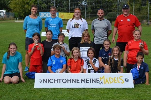 Gruppe von M&auml;dchen und Betreuern auf einem Sportplatz, mit Medaillen und einem Banner "#NICHTOHNEMEINEM&Auml;DLS".