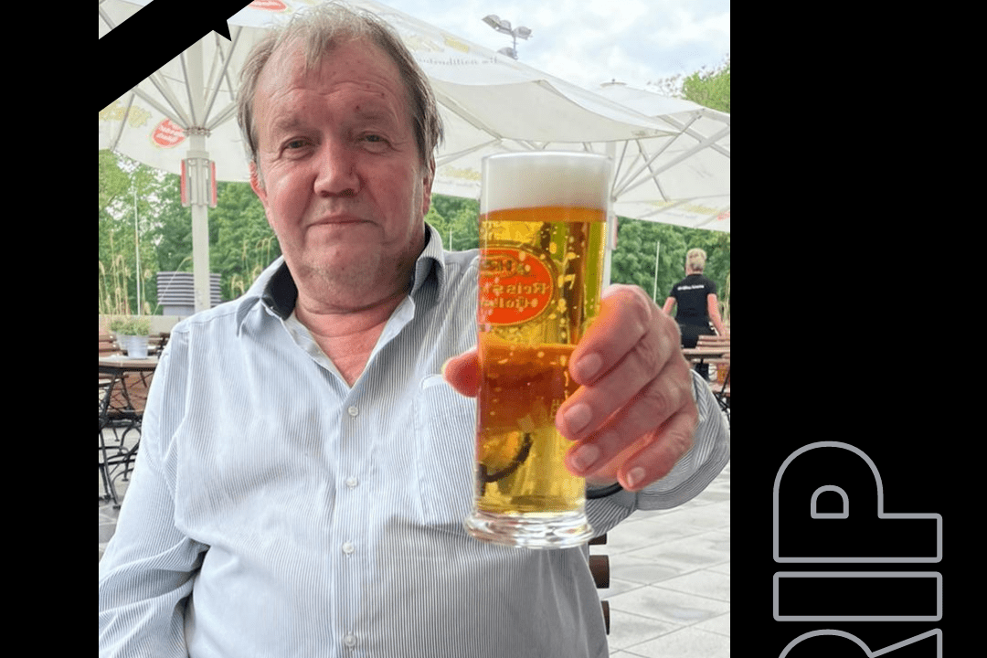 Mann h&auml;lt ein Glas Bier in der Hand, sitzt an einem Tisch im Freien unter Sonnenschirmen.