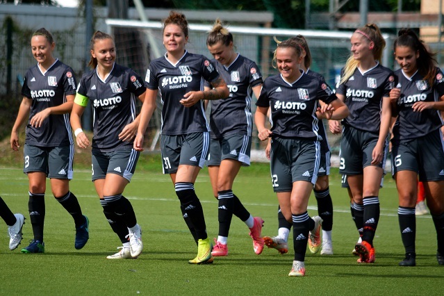 Gruppe von Fu&szlig;ballspielerinnen in grauen Trikots beim Laufen auf einem Fu&szlig;ballfeld unter Sonnenschein.
