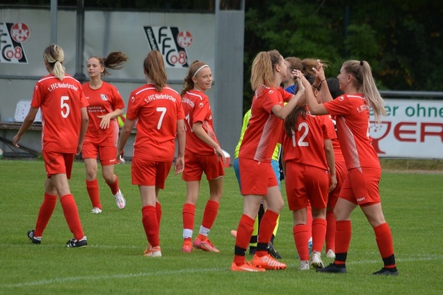 Mannschaft von Frauen in roten Trikots feiert Tor auf einem Fu&szlig;ballplatz mit gr&uuml;nem Rasen.
