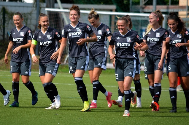 Gruppe von Fu&szlig;ballspielerinnen in grauen Trikots beim Laufen auf einem Fu&szlig;ballfeld unter Sonnenschein.