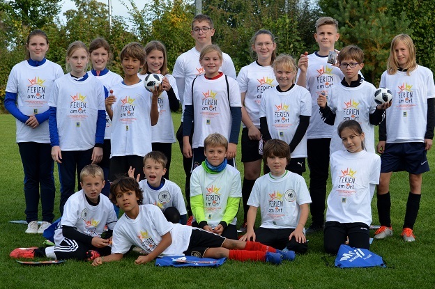 Gruppe von Kindern in wei&szlig;en T-Shirts mit Aufdruck, die auf einem Fu&szlig;ballfeld mit B&auml;llen posieren.