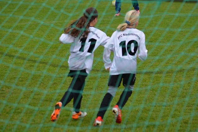 Zwei junge Fu&szlig;ballspielerinnen in wei&szlig;en Trikots mit den Nummern 11 und 10 laufen &uuml;ber das gr&uuml;ne Spielfeld.