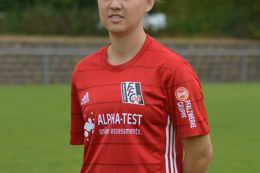 Junge Frau in rotem Fu&szlig;balltrikot steht auf einem Rasenplatz mit B&auml;umen im Hintergrund.