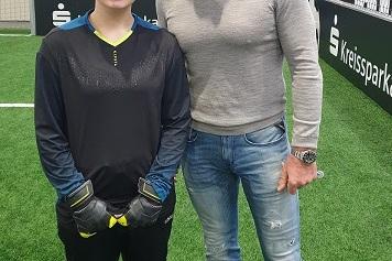 Zwei Personen stehen auf einem Sportplatz: eine junge Torh&uuml;terin in Trainingskleidung und ein Mann in Freizeitkleidung.