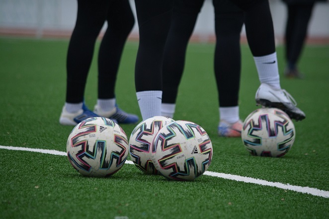 Nahaufnahme von drei Fu&szlig;b&auml;llen auf einem Kunstrasenplatz, umgeben von stehenden Spielern in Sportbekleidung.