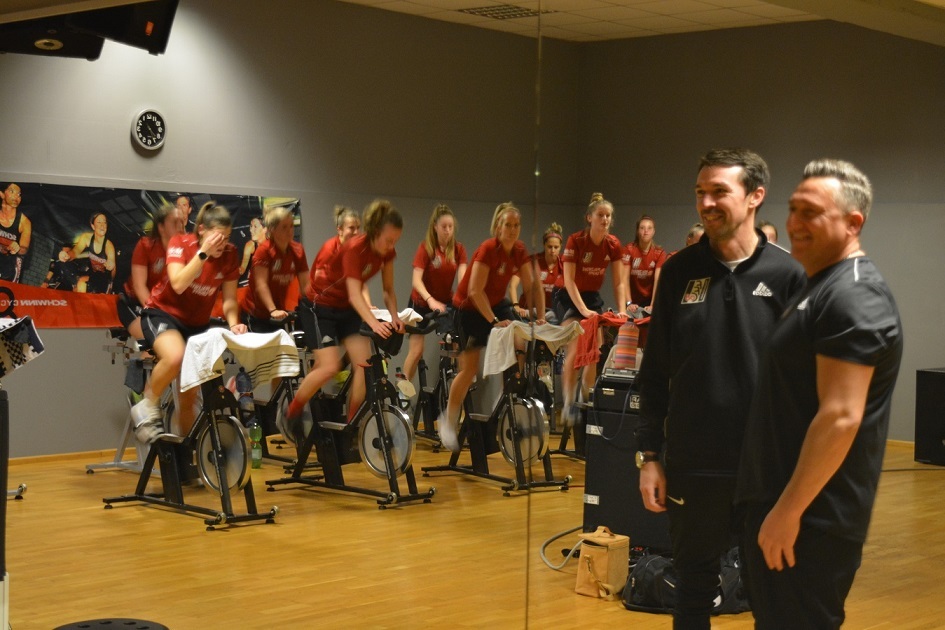 Zwei Trainer stehen l&auml;chelnd vor einer Gruppe weiblicher Sportlerinnen, die auf Fitnessbikes im Hintergrund trainieren.