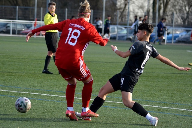 Zwei Fu&szlig;ballspielerinnen im Wettkampf um den Ball auf einem Rasenfeld, Schiedsrichter beobachtet die Szene.