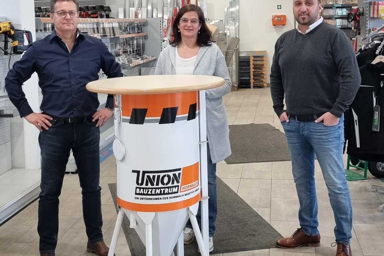 Drei Personen stehen an einem Stehtisch im Bauzentrum, im Hintergrund Werkzeuge und Bauzubehör sichtbar.