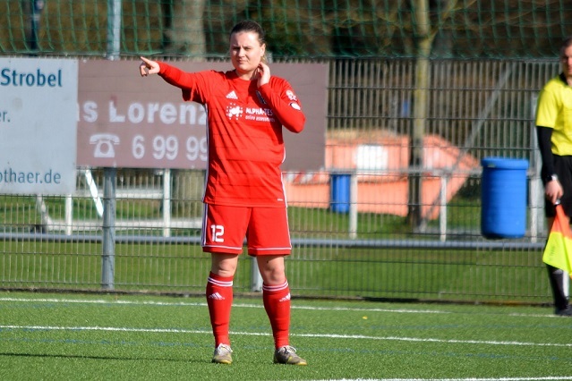 Spielerin in rotem Trikot zeigt mit dem Finger auf das Spielfeld, w&auml;hrend sie auf einem Fu&szlig;ballplatz steht.