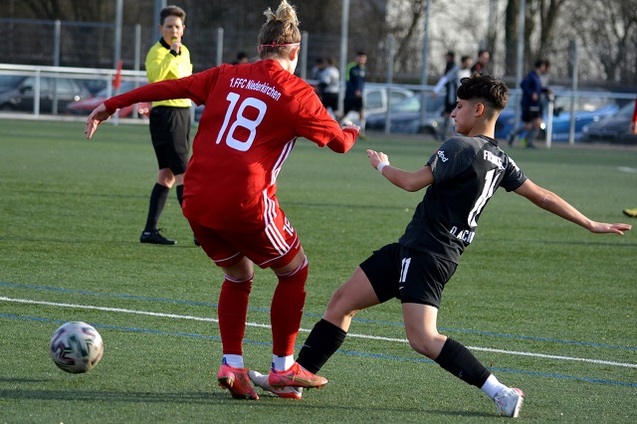 Zwei Fu&szlig;ballspielerinnen im Wettkampf um den Ball auf einem Rasenfeld, Schiedsrichter beobachtet die Szene.