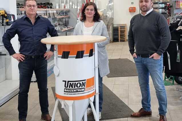 Drei Personen stehen an einem Stehtisch im Bauzentrum, im Hintergrund Werkzeuge und Bauzubeh&ouml;r sichtbar.