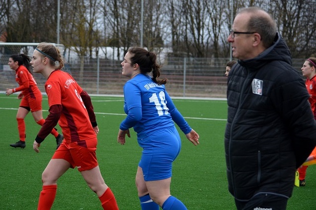 Zwei Fu&szlig;ballspielerinnen in roten und blauen Trikots bewegen sich auf einem Kunstrasenplatz, ein Trainer beobachtet das Spiel.