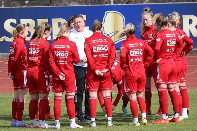 Trainer spricht mit einem Team in roten Trikots während einer Besprechung auf dem Fußballplatz.