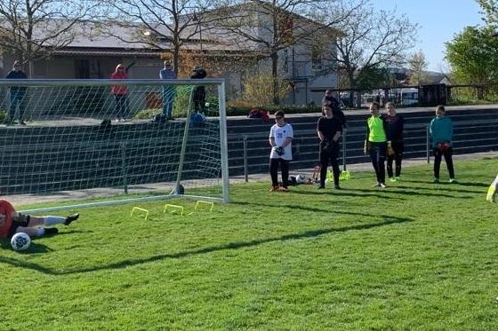 Torwart im Training, während ein Spieler einen Strafstoß auf das Tor schießt; Zuschauer beobachten am Rande des Spielfelds.