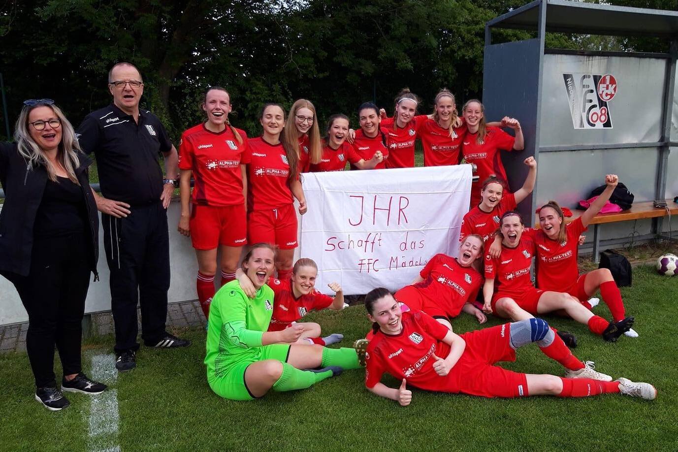 Mannschaft von Frauenfu&szlig;ballspielerinnen in roten Trikots posiert mit Banner nach einem Spiel am Fu&szlig;ballplatz.