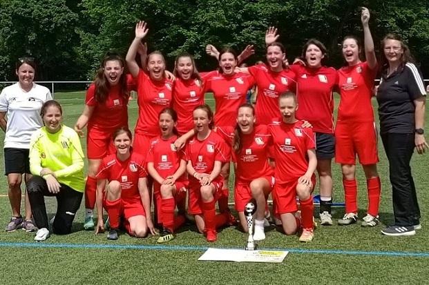 M&auml;dchenfu&szlig;ballmannschaft in roten Trikots posiert fr&ouml;hlich auf einem Sportplatz mit Pokal und Urkunde.