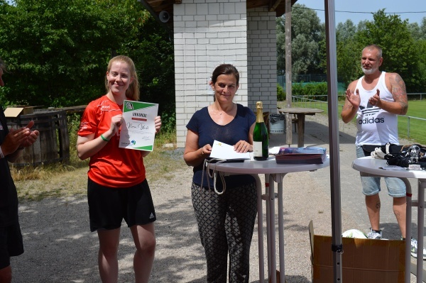 Junge Frau in rotem Trikot h&auml;lt Urkunde, w&auml;hrend eine andere Frau im Hintergrund mit Sektflasche am Tisch steht.