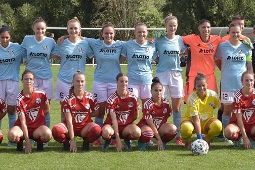 Zwei Frauenfu&szlig;ballmannschaften sitzen und stehen auf einem Sportfeld, alle in Trikots, mit Torwart in gelber Kleidung.