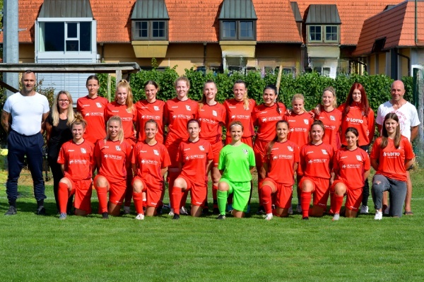 Gruppenportr&auml;t einer Frauenfu&szlig;ballmannschaft in roten Trikots, auf einem Rasenplatz aufgestellt, mit Trainer und Betreuern.