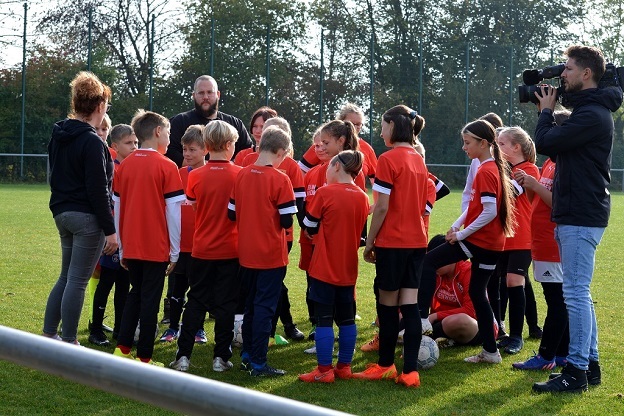 Eine Gruppe von Kindern in roten Fu&szlig;balltrikots versammelt sich um einen Trainer auf einem Spielfeld.
