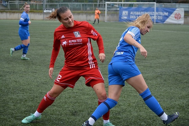 Zwei Spielerinnen im Zweikampf auf einem Fu&szlig;ballfeld, eine in rotem und die andere in blauem Trikot.