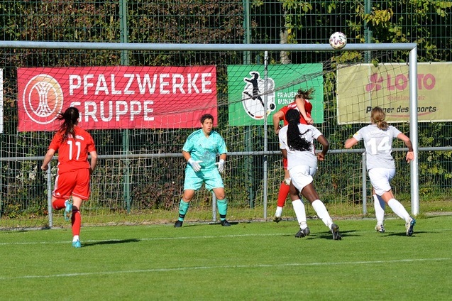 Spielszene eines Fu&szlig;ballspiels: Spielerin in Rot schie&szlig;t auf das Tor, Torwart in Gr&uuml;n springt zur Abwehr, Werbung im Hintergrund.