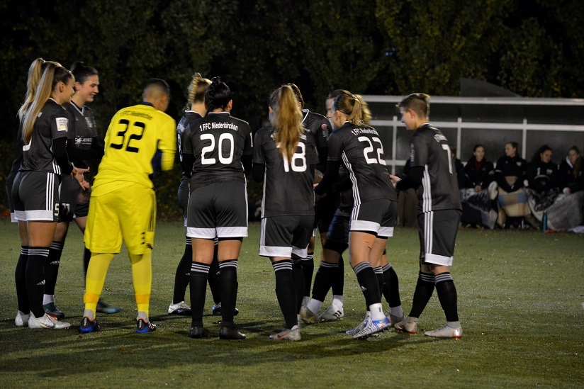 Eine Gruppe von Fu&szlig;ballspielerinnen in schwarzen Trikots versammelt sich auf dem Platz, um sich strategisch zu besprechen.