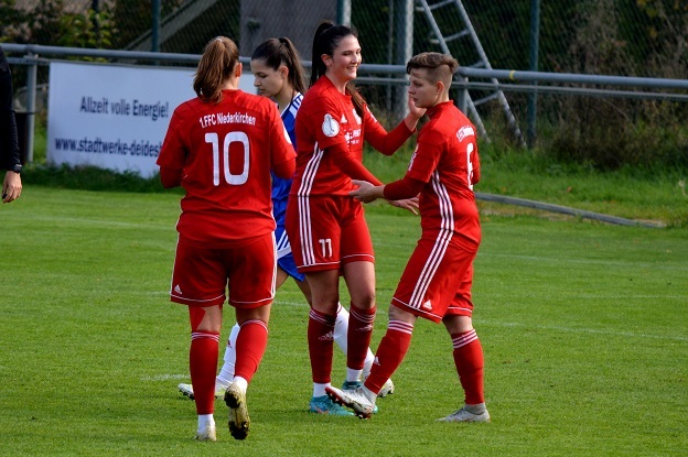 Vier Fu&szlig;ballspielerinnen in roten Trikots feiern auf einem gr&uuml;nen Platz, eine tr&auml;gt ein blaues Torwarttrikot.
